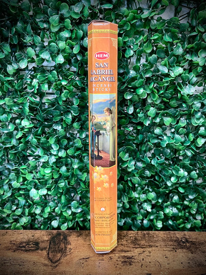 Hem San Abril Incense stick packaging on a wooden surface with green foliage in the background